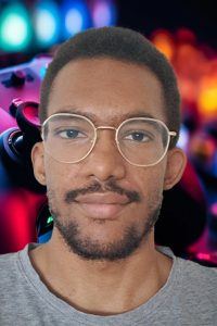 Portrait d’un jeune homme photographié de face, cadré du cou jusqu’au haut du buste. Il a les cheveux courts, porte des lunettes, a une barbe clairsemée, sourit légèrement et a les iris noirs.